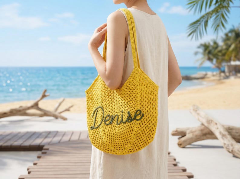 Pu&ograve; includere: Una borsa tote gialla all'uncinetto con il nome "Denise" ricamato in verde. La borsa ha una tracolla ed &egrave; tenuta da una persona. Lo sfondo mostra una scena di spiaggia con una passerella di legno e l'oceano.