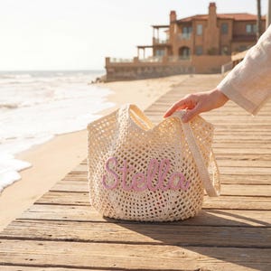 Pu&ograve; includere: Una borsa tote color crema a trama aperta con il nome "Stella" ricamato in rosa. La borsa ha due manici ed &egrave; tenuta da una persona su una passerella di legno vicino alla spiaggia. Lo sfondo comprende l'oceano e una casa.