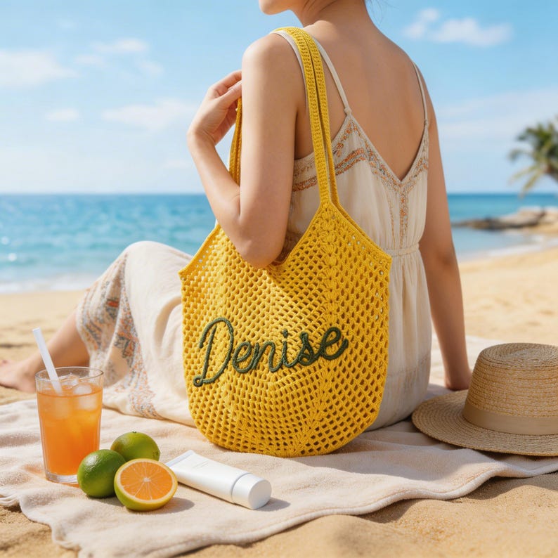 Pu&ograve; includere: Una borsa tote gialla all'uncinetto con il nome "Denise" ricamato in verde. La borsa ha lunghe cinghie e un design a rete. La borsa &egrave; su una spiaggia con una bevanda, frutta e un cappello.