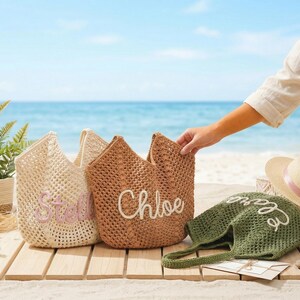 Pu&ograve; includere: Tre borse tote intrecciate in crema, marrone e verde oliva, ciascuna con un nome ricamato in un colore a contrasto. Un cappello di paglia con un nastro rosa &egrave; vicino. Le borse sono su una superficie di legno con una spiaggia e un oceano sullo sfondo.