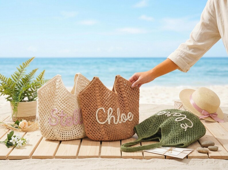 Pu&ograve; includere: Tre borse tote intrecciate in crema, marrone e verde oliva, ciascuna con un nome ricamato in un colore a contrasto. Un cappello di paglia con un nastro rosa &egrave; vicino. Le borse sono su una superficie di legno con una spiaggia e un oceano sullo sfondo.