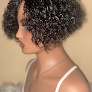 Pode incluir: Uma peruca curta de cor castanho-escuro com textura encaracolada, estilizada num corte bob em camadas. O cabelo apresenta caracóis definidos e apertados que emolduram o rosto, criando uma aparência volumosa. A peruca está num manequim.