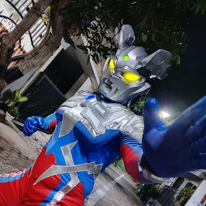 K&ouml;nnte beinhalten: Ein Superhelden-Kost&uuml;m mit einem silbernen Helm mit gelben Augen und einem blau-roten Body. Das Kost&uuml;m hat silberne Akzente und ein blauer Handschuh ist ausgestreckt. Das Design ist futuristisch und auff&auml;llig.