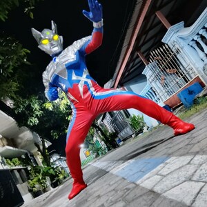 K&ouml;nnte beinhalten: Ein rot-blau-silberner Superheldenanzug mit silbernem Helm und gelben Augen. Der Anzug hat ein sternf&ouml;rmiges Design auf der Brust und blaue Handschuhe. Die Person tr&auml;gt rote Stiefel und posiert auf einer gepflasterten Oberfl&auml;che.