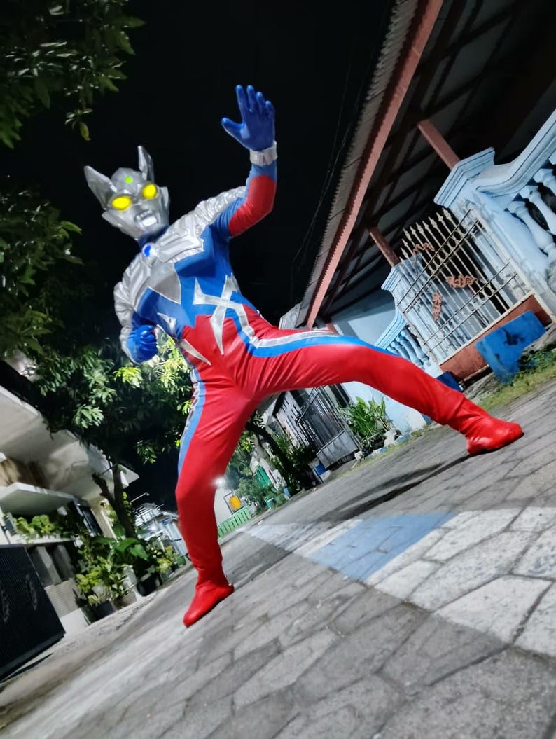 K&ouml;nnte beinhalten: Ein rot-blau-silberner Superheldenanzug mit silbernem Helm und gelben Augen. Der Anzug hat ein sternf&ouml;rmiges Design auf der Brust und blaue Handschuhe. Die Person tr&auml;gt rote Stiefel und posiert auf einer gepflasterten Oberfl&auml;che.