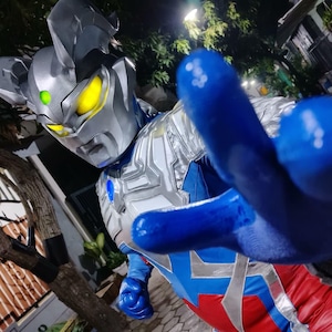 K&ouml;nnte beinhalten: Nahaufnahme eines Superhelden-Kost&uuml;ms. Der silberne Helm hat gelbe Augen und einen gr&uuml;nen Edelstein. Der Anzug kombiniert Blau, Rot und Silber. Die blaue, behandschuhte Hand des Superhelden ist auf den Betrachter gerichtet.