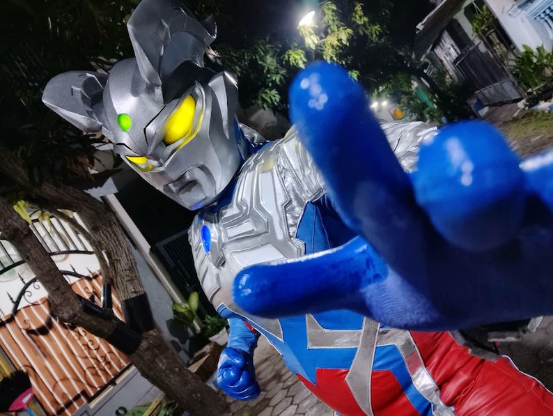 K&ouml;nnte beinhalten: Nahaufnahme eines Superhelden-Kost&uuml;ms. Der silberne Helm hat gelbe Augen und einen gr&uuml;nen Edelstein. Der Anzug kombiniert Blau, Rot und Silber. Die blaue, behandschuhte Hand des Superhelden ist auf den Betrachter gerichtet.