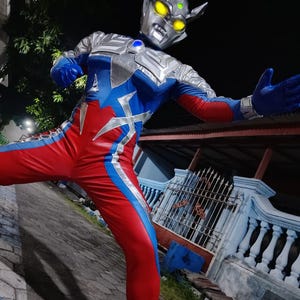 K&ouml;nnte beinhalten: Ein Ganzk&ouml;rperkost&uuml;m in Silber, Blau und Rot. Das Kost&uuml;m beinhaltet einen detaillierten Helm mit gelben Augen und einem blauen Edelstein. Der Anzug hat eine dynamische Pose, mit ausgestreckten Armen, die Action suggeriert.