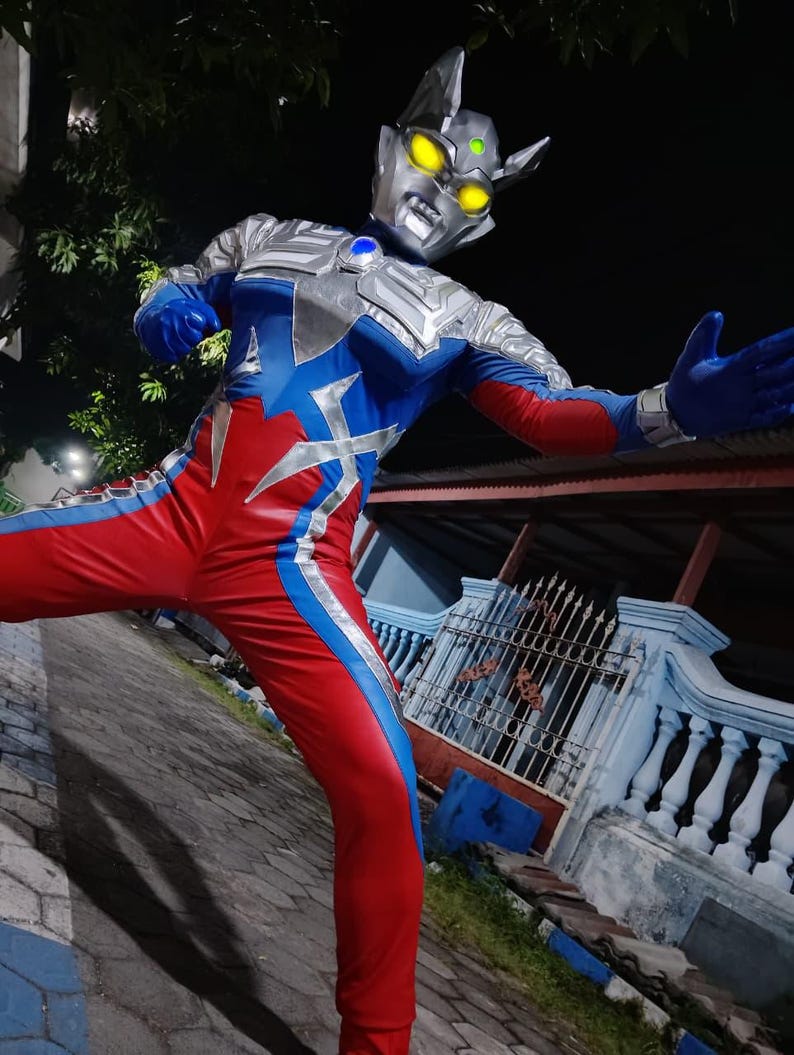 K&ouml;nnte beinhalten: Ein Ganzk&ouml;rperkost&uuml;m in Silber, Blau und Rot. Das Kost&uuml;m beinhaltet einen detaillierten Helm mit gelben Augen und einem blauen Edelstein. Der Anzug hat eine dynamische Pose, mit ausgestreckten Armen, die Action suggeriert.