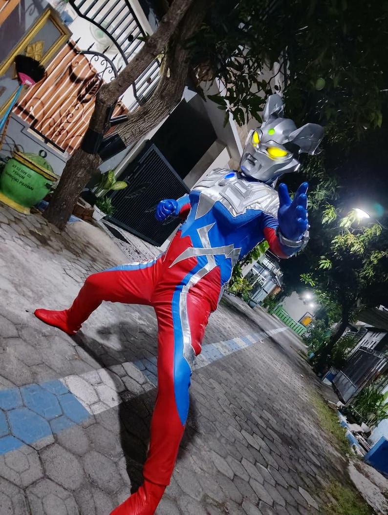 K&ouml;nnte beinhalten: Ein Ganzk&ouml;rperkost&uuml;m mit einem silbernen Helm mit gelben Augen, einem blau-roten Body mit silbernen Akzenten und roten Stiefeln. Das Kost&uuml;m ist so gestaltet, dass es einem Superhelden-Charakter &auml;hnelt.