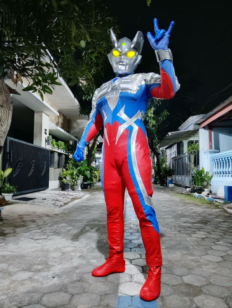 K&ouml;nnte beinhalten: Ein rot, blau und silberfarbenes Superheldenkost&uuml;m. Das Kost&uuml;m hat einen silbernen Helm mit gelben Augen und eine blaue Handgeste. Das Outfit umfasst rote Stiefel und Handschuhe.