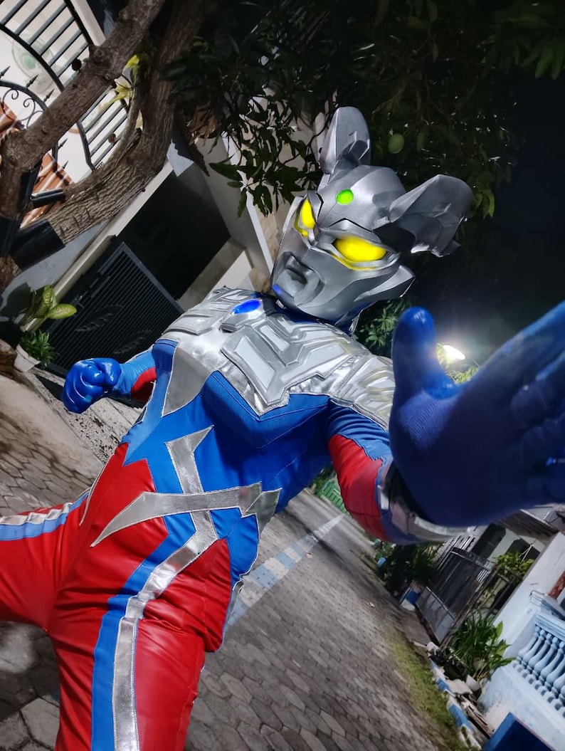 K&ouml;nnte beinhalten: Ein Superhelden-Kost&uuml;m mit einem silbernen Helm mit gelben Augen und einem blau-roten Body. Das Kost&uuml;m hat silberne Akzente und ein blauer Handschuh ist ausgestreckt. Das Design ist futuristisch und auff&auml;llig.