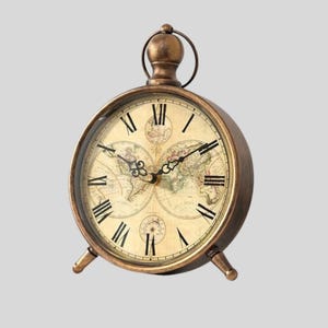 Puede incluir: Reloj de escritorio de estilo antiguo con acabado bronce. La esfera del reloj presenta un diseño de mapa del mundo con números romanos. El reloj tiene forma redonda, soportado por dos pequeños pies y un lazo en la parte superior.