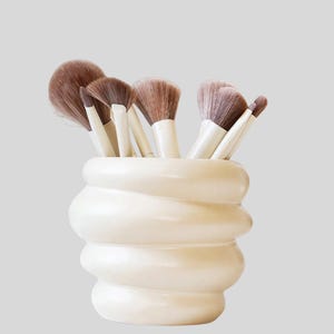 Op de afbeelding: Een verzameling make-up borstels met beige handvatten en bruine borstelharen is gerangschikt in een crèmekleurige, gedraaide keramische houder. De houder heeft een uniek, gestapeld ontwerp en de borstels zijn klaar voor gebruik.