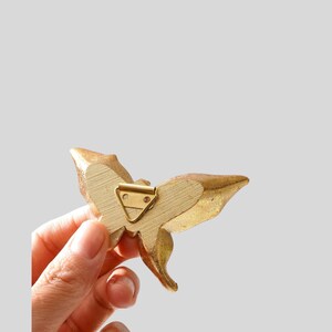 May include: A gold-colored butterfly-shaped decorative object with a metal clip on the back. The butterfly has detailed wing patterns and is held in a person's hand against a light gray background.