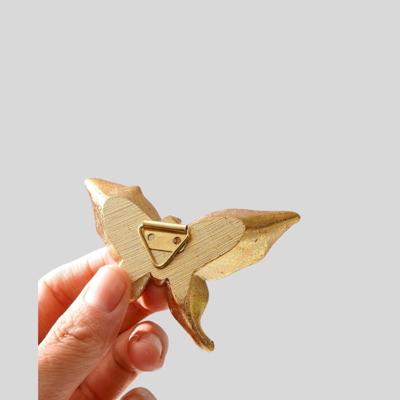 May include: A gold-colored butterfly-shaped decorative object with a metal clip on the back. The butterfly has detailed wing patterns and is held in a person's hand against a light gray background.