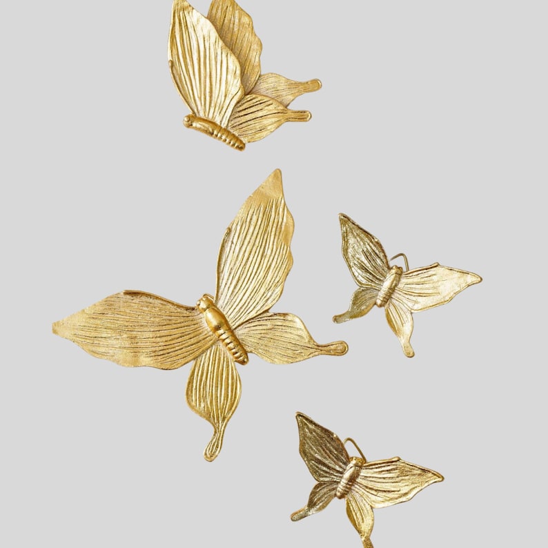 May include: Four gold-colored butterfly wall decorations. The butterflies have detailed wing patterns and are arranged in a visually appealing composition. The butterflies are made of a metallic material and are set against a neutral background.