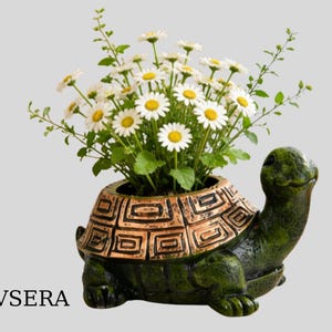 Puede incluir: Una maceta verde con forma de tortuga con un diseño de caparazón de color bronce, llena de margaritas blancas y follaje verde. La maceta tiene un acabado texturizado y es adecuada para uso en interiores o exteriores.
