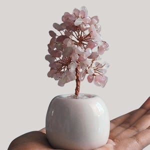 Puede incluir: Una escultura decorativa de árbol con un tronco y ramas de alambre de cobre, adornada con hojas de piedras preciosas rosas. El árbol está en una pequeña maceta de cerámica blanca y redondeada. La escultura está en la mano de una persona.