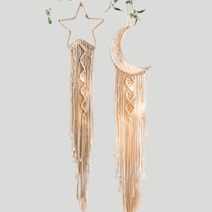 May include: Two macrame wall hangings, one shaped like a star and the other a crescent moon. Both are made of cream-colored woven cotton and feature intricate knotting patterns. The star and moon have hanging strands and are illuminated with soft lights.