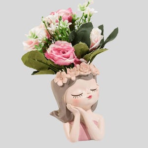 Puede incluir: Un macetero decorativo con forma de cabeza de niña, con los ojos cerrados y una expresión serena. El macetero está lleno de rosas rosas y blancas, hojas verdes y pequeñas flores blancas. La cabeza de la niña está adornada con una corona floral.