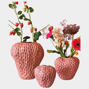 Puede incluir: Tres jarrones rosas de cerámica con forma de fresa de diferentes tamaños. Los jarrones tienen una superficie texturizada con pequeñas hendiduras. Los jarrones están llenos de flores y ramas de fresa.