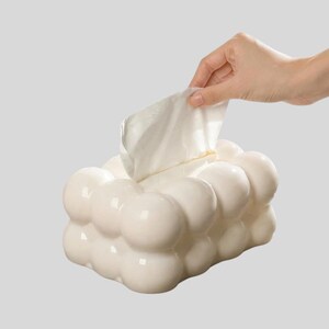 Puede incluir: Un dispensador de pañuelos blanco con forma de burbujas, con una mano sacando un pañuelo. El dispensador tiene un acabado brillante y un diseño moderno único. Los pañuelos son blancos y plegados.