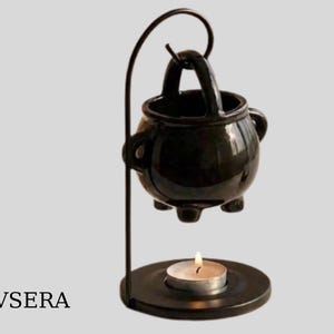Peut inclure: Un chauffe-huile en forme de chaudron en céramique noire avec une poignée, soutenu par un support en métal noir. Une bougie chauffe-plat allumée est posée sur une base ronde sous le chaudron. Le mot "EVSERA" est en bas à gauche.