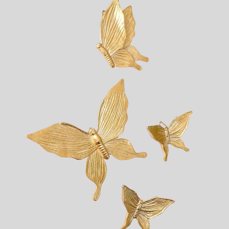 May include: Four gold-colored butterfly wall decorations. The butterflies are in various sizes and poses, with detailed wing textures. The background is a solid, light gray color. The butterflies appear to be made of metal.