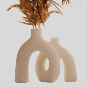 May include: A beige ceramic vase with an abstract design, featuring an arched shape and a circular opening. The vase holds dried, feathery brown plants. The neutral color and unique shape make it a decorative home accent.
