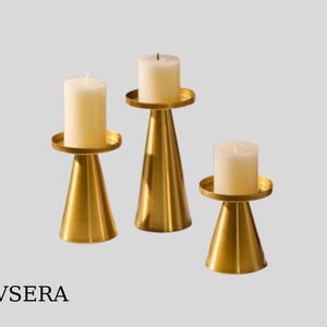 Puede incluir: Tres candelabros dorados con base cónica y parte superior redonda. Cada uno sostiene una vela de pilar blanca. Los candelabros varían en altura, creando una disposición visualmente atractiva. La marca "EVSERA" está en la parte inferior.