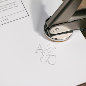 Könnte beinhalten: Eine silberne Metall-Siegelpresse, die die Initialen "A & C" auf weißes Papier stempelt. Das Wort "DREAMING" ist in der oberen linken Ecke sichtbar. Das Papier liegt auf einer braunen Oberfläche.