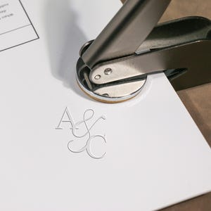Op de afbeelding: Een zilverkleurige metalen embossermachine die initialen op wit papier drukt. De in reliëf aangebrachte initialen zijn "A&C". De embossermachine staat schuin, met het papier op een lichtbruin oppervlak.