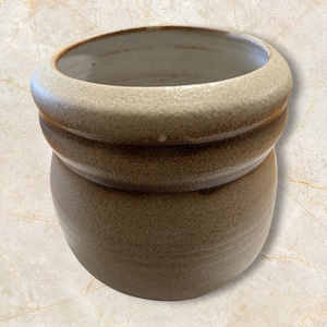 May include: A round ceramic pot with a thick, rounded rim and a textured, speckled tan exterior. The interior of the vessel is smooth and white, contrasting with the earthy, matte finish of the outer surface. The pot has a wide, open top.