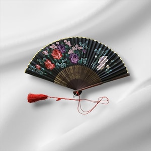 May include: A black hand fan with a floral design featuring red and purple roses, accented with gold trim. A red tassel hangs from the base of the fan. The fan is open, displaying its full, semi-circular shape.