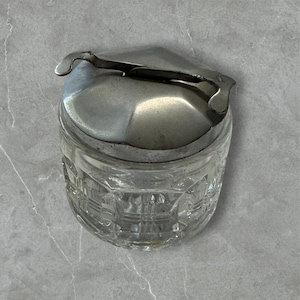 May include: A clear glass jar with a silver metal lid and handle. The jar has a geometric pattern on the sides. The lid is slightly domed and has a curved handle for easy opening. The jar is likely for storing sugar or other small items.