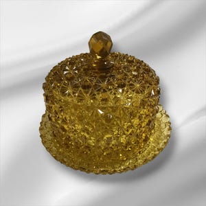 May include: A vintage amber-colored glass jar with a lid. The jar and lid feature a diamond-cut pattern, and the lid is topped with a faceted knob. The jar sits on a matching saucer, creating a cohesive set. This decorative piece is suitable for storing small items.