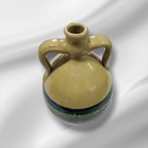 May include: A small, yellow ceramic jug with two handles and a rounded body. The jug features a dark blue and green striped band around the base. The jug is likely handmade and has a rustic, artisanal aesthetic.