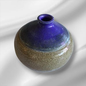 May include: A handcrafted ceramic vase with a rounded base and a narrow neck. The upper portion is a deep blue, while the lower half is a speckled, earthy brown. The vase has a glossy finish and a unique, organic shape.