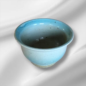 May include: A handmade ceramic bowl with a light blue glaze on the rim and a speckled, off-white body. The bowl has a rounded shape and a slightly flared rim. The base is a natural, unglazed clay color.