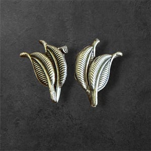 May include: A pair of silver-toned leaf-shaped earrings. Each earring features three detailed leaves with textured veins, creating a naturalistic design. The earrings are set against a dark gray background.