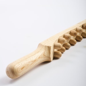 May include: A light-colored wooden massage tool with a rounded handle and a series of rounded knobs along its length. The tool is designed for body massage and is made of natural wood.