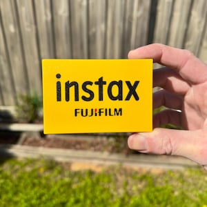 Peut inclure: Un produit rectangulaire jaune avec le mot "instax" en noir et "FUJIFILM" en dessous. Le produit est tenu dans la main d'une personne, avec une clôture en bois et de l'herbe verte en arrière-plan.