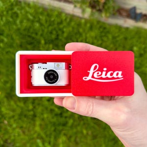 Può includere: Una fotocamera digitale in miniatura color argento con un obiettivo nero che riporta la scritta 2560x1440 f=2.0mm F2.4, alloggiata in una scatola bianca con coperchio scorrevole rosso che mostra il logo Leica bianco. La fotocamera è in un vano rosso.