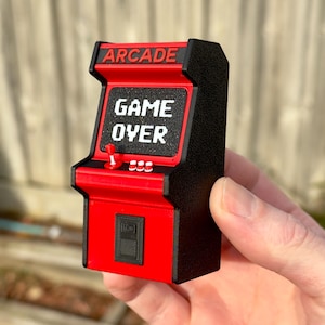 May include: A miniature arcade cabinet model in black and red with a textured finish. The screen displays the white text GAME OVER, while the top marquee reads ARCADE. It features a red joystick, three white buttons, and a black coin slot panel.