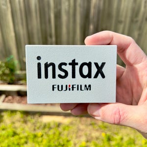 Op de afbeelding: Een rechthoekige, lichtgrijze doos met het woord "instax" in grote, zwarte, vette letters. Daaronder staat het woord "FUJIFILM" in kleinere, zwarte letters. Een kleine rode stip staat tussen de "x" en de "F".