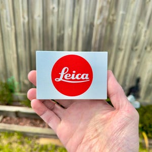 Puede incluir: Un objeto rectangular gris claro con un círculo rojo con la palabra "Leica" en blanco. El objeto se sostiene en una mano. El fondo es una valla de madera borrosa y vegetación.