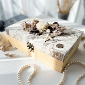 May include: A rectangular wooden box with a marbled lid, decorated with seashells, a starfish, and dried flowers. The box has a light wood base and a decorative clasp. A pearl necklace is in the foreground.