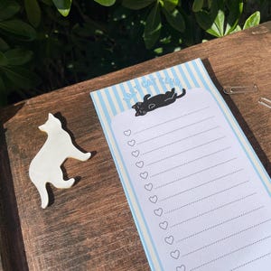 Può includere: Una superficie in legno ospita un magnete a forma di gatto bianco e un blocco note con strisce blu e la scritta "LAZY CAT CLUB". Il blocco note presenta un'illustrazione di gatto nero e punti elenco a forma di cuore. Sono presenti anche due graffette argentate.