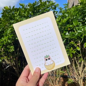 May include: A notepad with a pale yellow border and a white interior featuring a dotted line pattern. A cartoon cat wearing sunglasses and a pineapple hat is in a pineapple basket. The notepad is held in a person's hand.